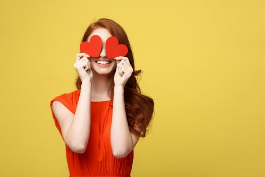 Lifestyle Concept: Attractive woman with beaming smile having two small red hearts in hands, closing eyes with paper heart symbols while standing over yellow background