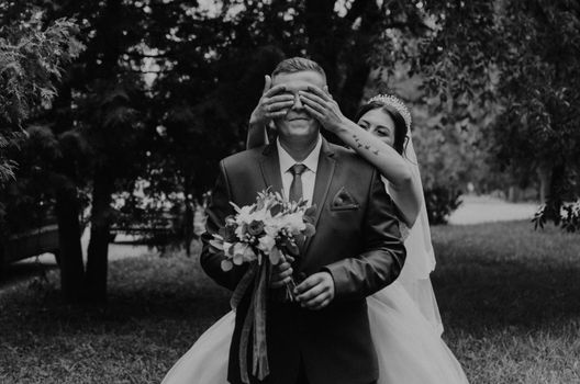 European Caucasian man groom in suit and woman bride in wedding dress veil tiara on head. first meeting newlyweds groom wait sees bride. girl comes up from behind closes guy's eyes with her hands