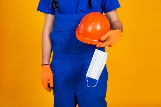 Worker in construction overalls and gloves on a yellow background, holds a protective helmet and a medical mask in his hand. The concept of the economic crisis, unemployment production, coronavirus.
