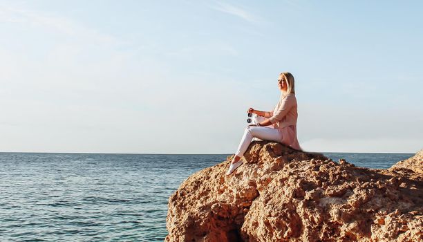 Blonde girl in sunglasses sits on a stone near the sea. A woman in pink clothes and sunglasses looks at the sea, enjoys the rest on a trip.