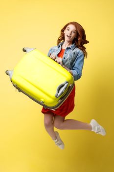 Travel and Lifestyle Concept: Portrait of a beautiful red hair fashion woman jumping and holding a bright green suitcase isolated over bright yellow background