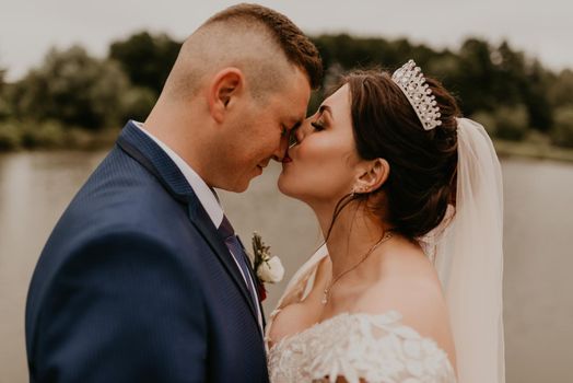 blonde European Caucasian young man groom in blue suit and black-haired woman bride in white wedding dress with long veil and tiara on head. Newlyweds hold hands smile kiss and look at each other near river