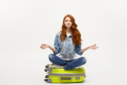 Traveling concept. Young pretty ginger woman meditating in lotus pose on the luggage valise. Isolated on white