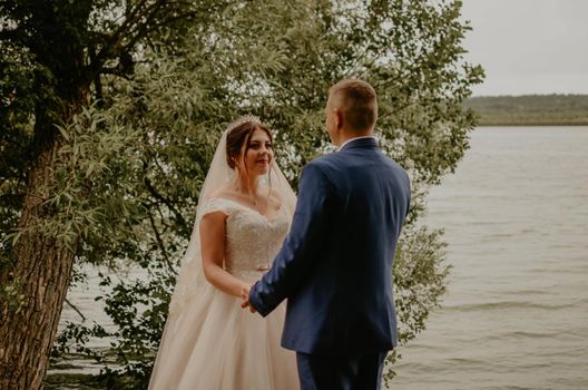 blonde European Caucasian young man groom in blue suit and black-haired woman bride in white wedding dress with long veil and tiara on head. Newlyweds hold hands smile kiss and look at each other near river