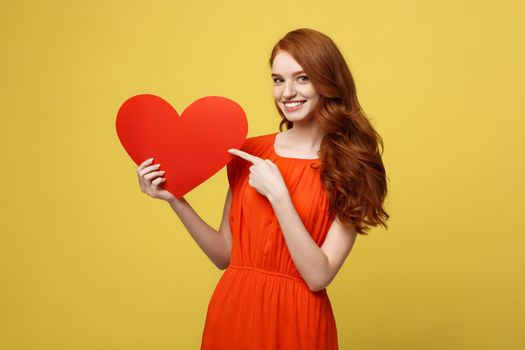 Valentine and Lifestyle Concept: Portrait of an attractive young woman dressed in red dress pointing finger at paper heart isolated over yellow studio background