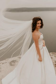 young brunette bride woman with short haircut in white wedding corset dress strapless and with a long chiffon veil on head. natural light nude makeup. Cool autumn weather. background large lake river