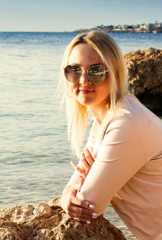 Blonde girl in sunglasses sits on a stone near the sea. A woman in pink clothes and sunglasses looks at the sea, enjoys the rest on a trip.