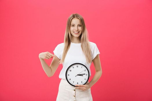 Portrait of an excited young girl dressed in white t-shirt pointing at alarm clock and looking at camera isolated over pink background.