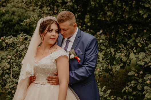 blonde European Caucasian young man groom in blue suit and black-haired woman bride in white wedding dress with long veil and tiara on head. newlyweds guy hugs a girl from behind