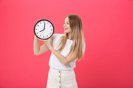 Portrait of an excited young girl dressed in white t-shirt pointing at alarm clock and looking at camera isolated over pink background.