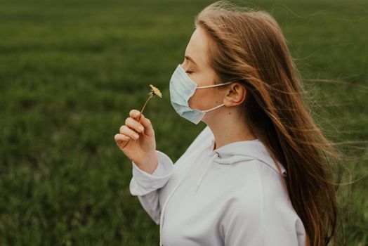 A girl in a respirator sniffs a yellow dandelion. Young blond European woman in a protective mask outdoors.