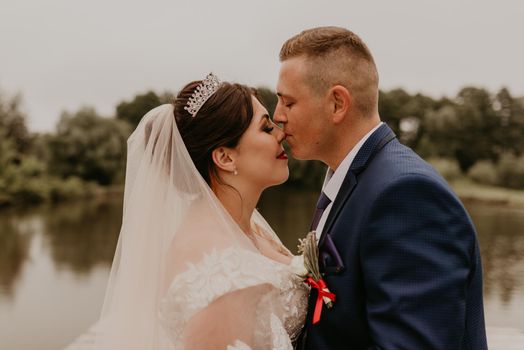 blonde European Caucasian young man groom in blue suit and black-haired woman bride in white wedding dress with long veil and tiara on head. Newlyweds hold hands smile kiss and look at each other near river