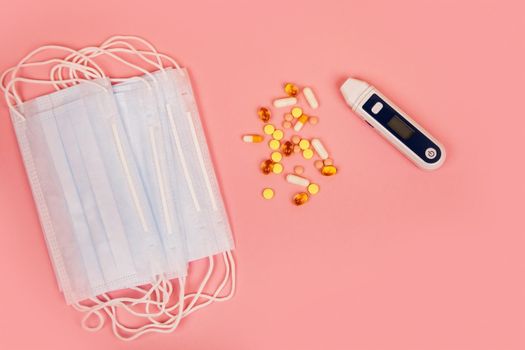 Medical disposable masks, pills and electronic thermometer on a pink background. Protection against influenza virus and pneumonia. Healthcare and medical concept.