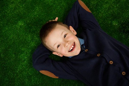 A little boy lies on his back, looking at the camera and smiling.. Top view of a cute happy little boy looking at camera and smiling while lies on a lawn