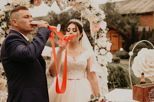 Bride and groom in wedding dress veil at wedding ceremony on flower arch. Newlyweds drink champagne with glasses together with red ribbon tied together. Slavic Ukrainian Russian traditions