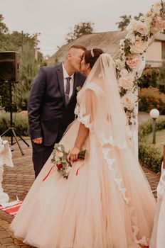 Bride and groom in a wedding dress and a long veil at a wedding ceremony on the background of a flower arch laughing kissing rejoicing. Slavic Ukrainian Russian traditions