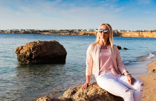 Blonde girl in sunglasses sits on a stone near the sea. A woman in pink clothes and sunglasses looks at the sea, enjoys the rest on a trip.