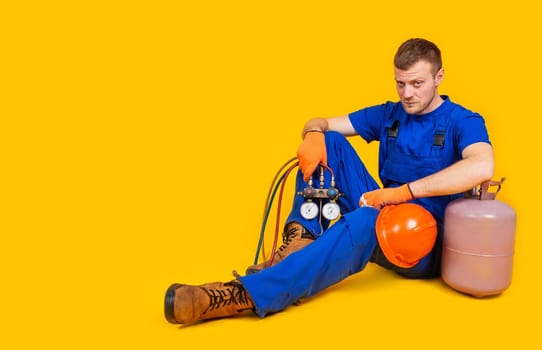 Sad fired worker with safety helmet sitting on yellow background. A man in overalls fired from work. The concept of the economic crisis, unemployment and production, coronavirus, pandemic, health.