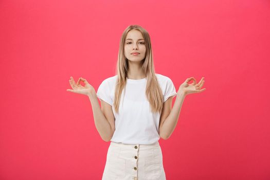 Blonde beauiful young female makes mudra sign, relaxes after hard working day, keeps eyes shut, practises yoga against pink background. Young pretty woman meditates indoor. Relaxation concept