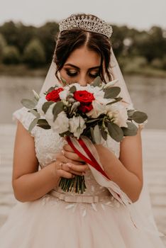European Caucasian young black-haired woman bride in white wedding dress with long veil and tiara on head. girl holding her bouquet flowers in hands. bride stands on wooden pier on background of river