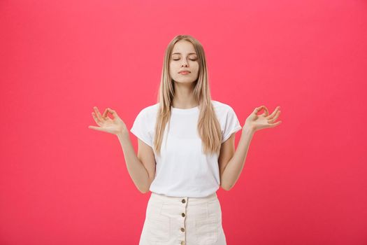Blonde beauiful young female makes mudra sign, relaxes after hard working day, keeps eyes shut, practises yoga against pink background. Young pretty woman meditates indoor. Relaxation concept