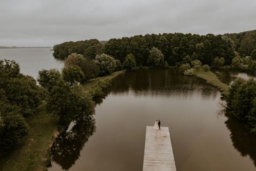 European Caucasian man groom in suit and black-haired woman bride in white wedding dress with long veil and tiara on head. Newlyweds kissing hugging on wooden pier bridge on lake. aerial view drone