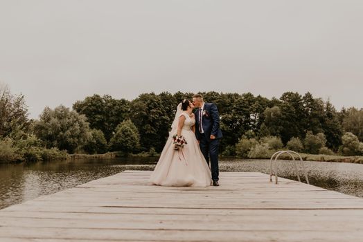 blonde European Caucasian young man groom in blue suit and black-haired woman bride in white wedding dress with long veil and tiara on head. Newlyweds kissing hugging on wooden pier bridge on lake