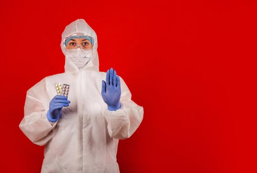 A doctor in a medical overalls shows a stop gesture. Young professional doctor woman holding pharmaceutical pills and hand making protection gesture sign with a serious and confident expression