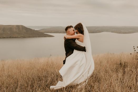 loving couple wedding newlyweds in white dress veil sports shoes and suit hug kissing whirl on tall grass in summer field on mountain above the river. sunset.