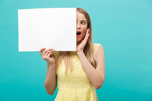 Portrait of amazed young blond woman holding blank sign with copy space on blue studio background. Showing shocked surprise face