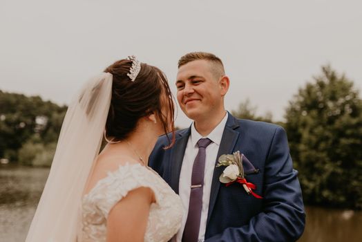 blonde European Caucasian young man groom in blue suit and black-haired woman bride in white wedding dress with long veil and tiara on head. Newlyweds hold hands smile kiss and look at each other near river