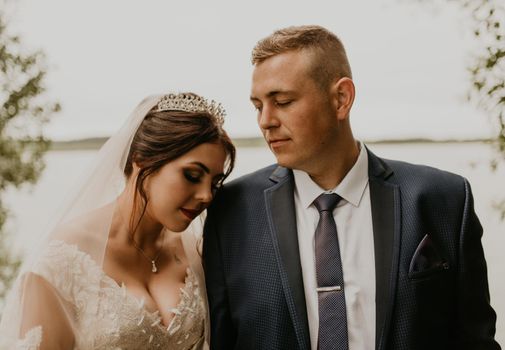 blonde European Caucasian young man groom in blue suit and black-haired woman bride in white wedding dress with long veil and tiara on head. Newlyweds hold hands smile kiss and look at each other near river