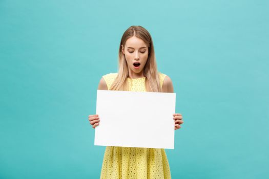 Closeup portrait, young woman in yellow dress holding white plain paper with shocked expression at what she sees, isolated blue background