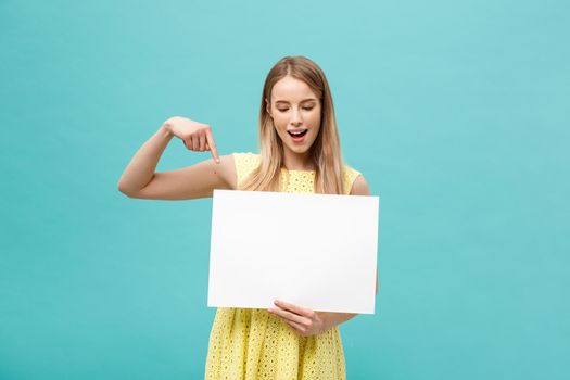 Portrait of young woman in yellow dress pointing finger at side white blank board. Isolated over Blue background