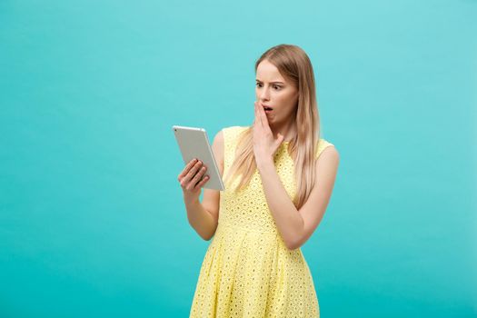 Portrait of a surprised amazed young woman looking at digital tablet isolated on a blue studio background