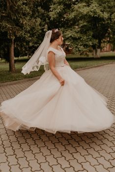 dark-haired European Caucasian young woman bride in white wedding dress with long veil and tiara on head spinning whirls dancing posing moving at nature on background greenery. Valentine love couple