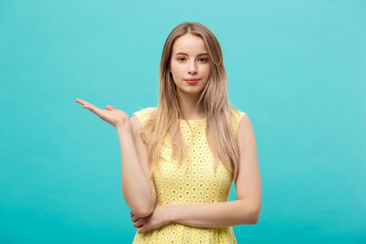 Portrait of stylish, nice, cute, caucasian woman with hairstyle in yellow dress holding in hand on empty places, copy spaces on her palms, looking at camera, isolated on blue background.