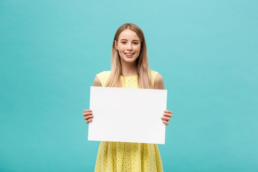 Lifestyle Concept: young beautiful girl smiling and holding a blank sheet of paper, dressed in yellow, isolated on pastel blue background.