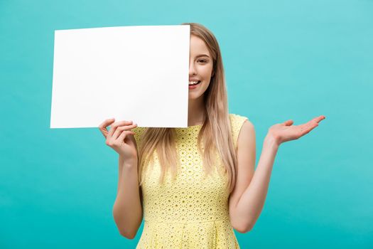 Lifestyle Concept: young beautiful girl smiling and holding a blank sheet of paper, dressed in yellow, isolated on pastel blue background.