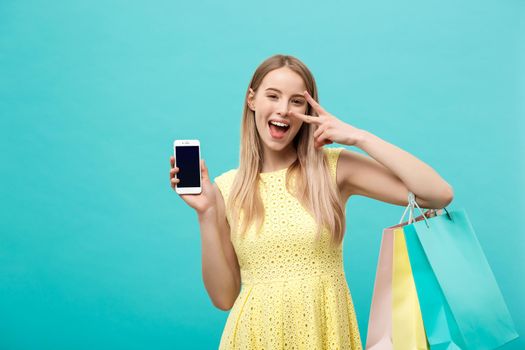 Portrait young attractive woman with shopping bags shows the phone's screen directly to the camera. Isolated on blue background.