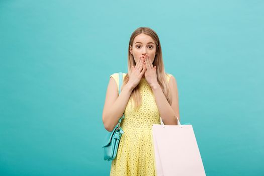 Lifestyle Concept: Portrait of shocked young attractive woman in yellow summer dressposing with shopping bags and looking at camera over blue background