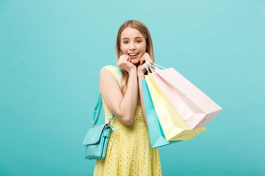 Lifestyle Concept: Portrait of shocked young attractive woman in yellow summer dressposing with shopping bags and looking at camera over blue background