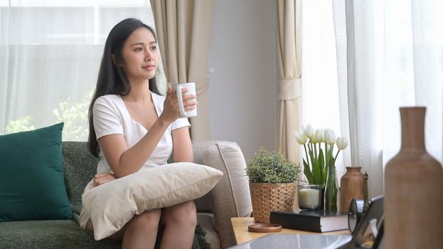 Relaxed millennial asian woman sitting on couch and drinking hot coffee in the morning.