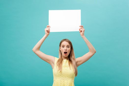 Closeup portrait, young woman in yellow dress holding white plain paper with shocked expression at what she sees, isolated blue background