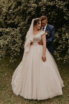 blonde European Caucasian young man groom in blue suit and black-haired woman bride in white wedding dress with long veil and tiara on head. newlyweds guy hugs a girl from behind