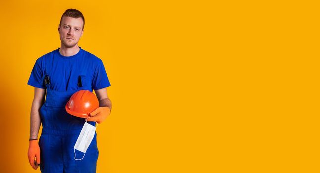Worker in construction overalls and gloves on a yellow background, holds a protective helmet and a medical mask in his hand. The concept of the economic crisis, unemployment production, coronavirus.