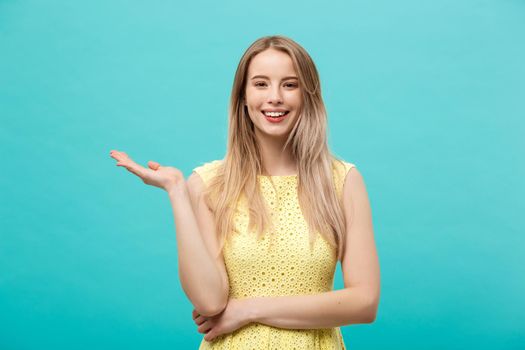 Portrait of stylish, nice, cute, caucasian woman with hairstyle in yellow dress holding in hand on empty places, copy spaces on her palms, looking at camera, isolated on blue background.