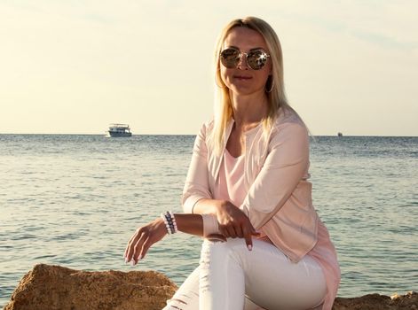 Blonde girl in sunglasses sits on a stone near the sea. A woman in pink clothes and sunglasses looks at the sea, enjoys the rest on a trip.