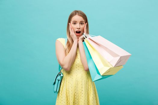 Lifestyle Concept: Portrait shocked young brunette woman in yellow summer dress posing with shopping bags isolated over pastel blue background.