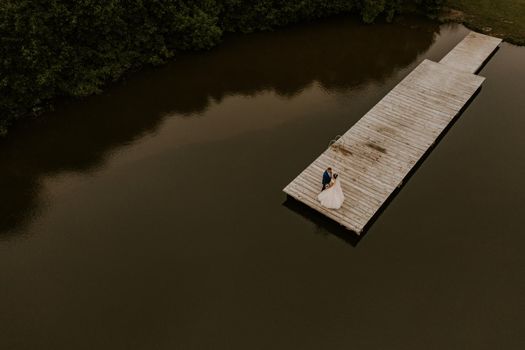 European Caucasian man groom in suit and black-haired woman bride in white wedding dress with long veil and tiara on head. Newlyweds kissing hugging on wooden pier bridge on lake. aerial view drone